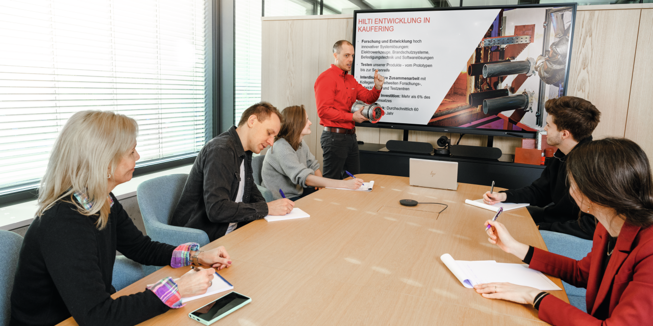Ein Bild zeigt eine Hilti-Schulungsveranstaltung, an der 5 Personen an einem Tisch teilnehmen und ein Hilti-Spezialist auf einem Bildschirm am Ende des Tisches Schulungsmaterialien vorführt