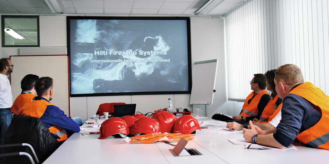 Ein Bild zeigt eine Hilti-Schulungsveranstaltung, an der 5 Personen an einem Tisch teilnehmen und ein Hilti-Spezialist auf einem Bildschirm am Tisch Schulungsmaterialien vorführt