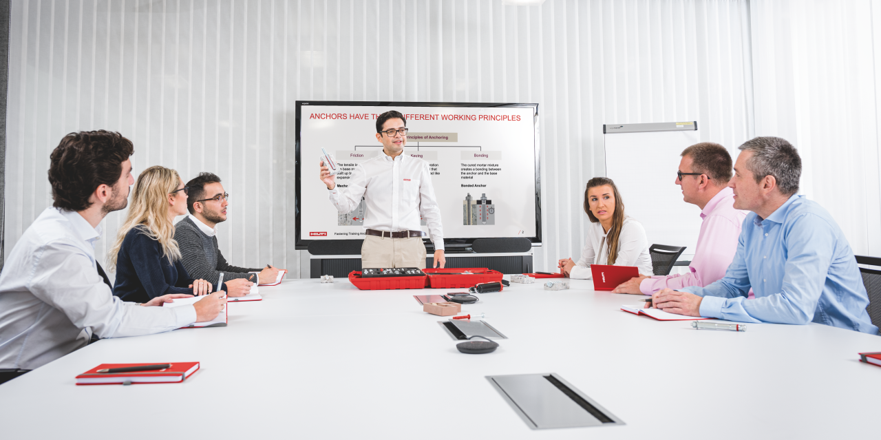 Bild einer Präsentation, bei der 4 Männer und 2 Frauen am Tisch sitzen und ein Hilti-Spezialist vor einem Bildschirm präsentiert.