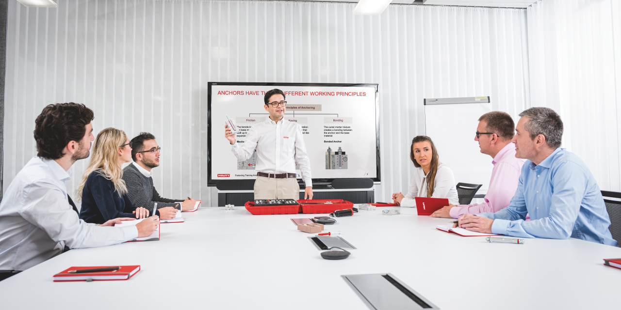 Bild einer Präsentation, bei der 4 Männer und 2 Frauen am Tisch sitzen und ein Hilti-Spezialist vor einem Bildschirm präsentiert.