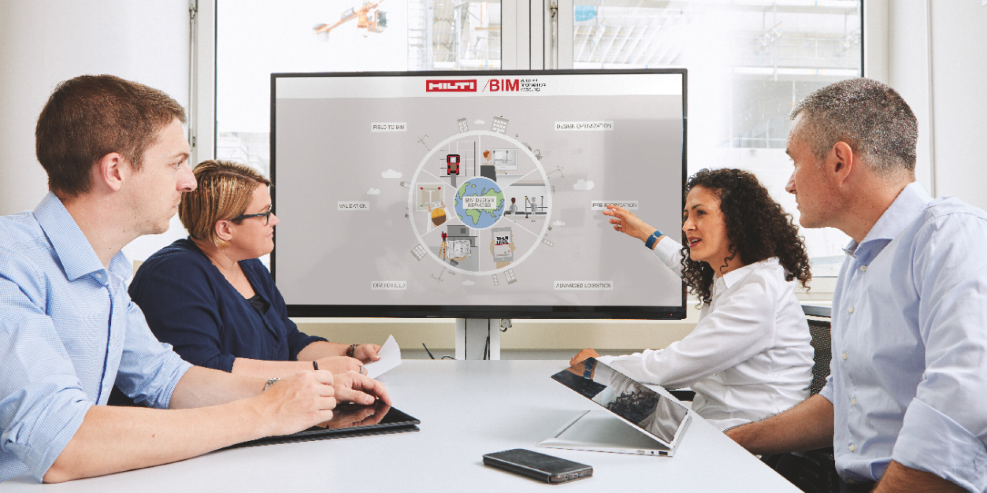 Das Bild zeigt vier Mitarbeiter, zwei Männer und zwei Frauen, die am Tisch sitzen, während eine der Frauen eine Präsentation über Hilti BIM hält. Die Präsentation wird auf dem Bildschirm angezeigt, der an einer Seite des Tisches steht.
