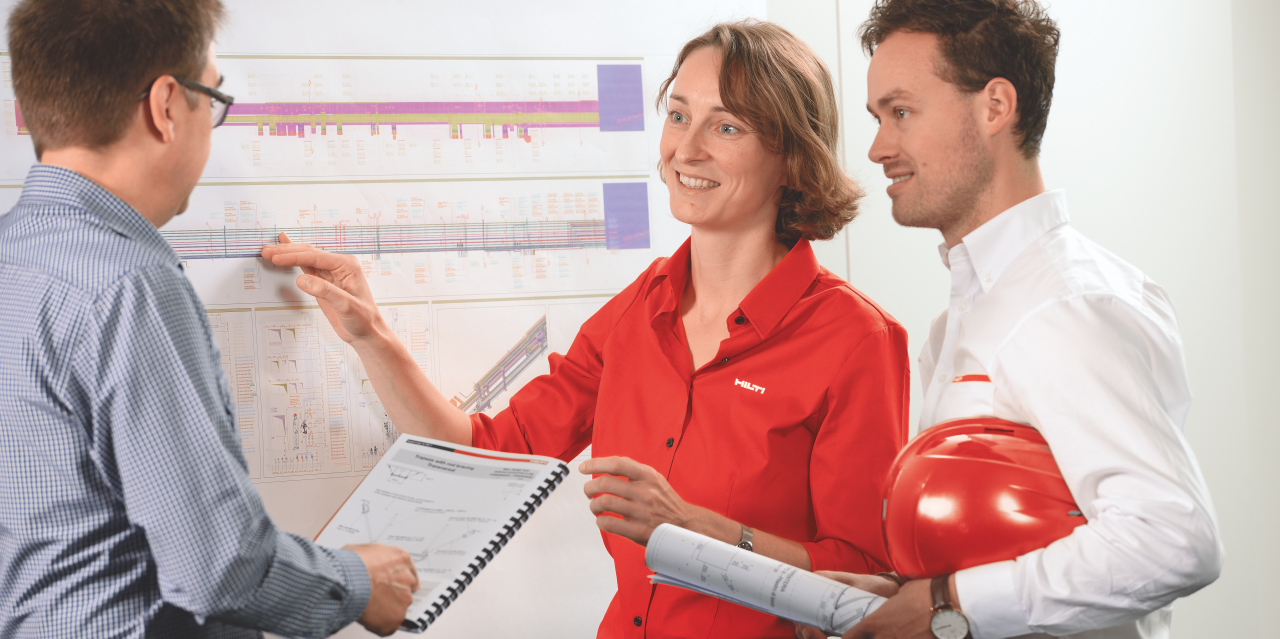 Bild mit zwei Hilti-Beratern, Frau und Mann, rechts und Kunde links. Sie stehen an der Wand mit dem Diagramm und unterhalten sich.